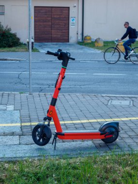 Cremona, İtalya - 27 Mayıs 2024 Kırmızı Kiralık Scooter arka planda bulanık bir bisikletçiyle kaldırıma park edilmiş, mikro hareketlilik, kentsel ulaşım ve sürdürülebilirlik gibi kavramlar