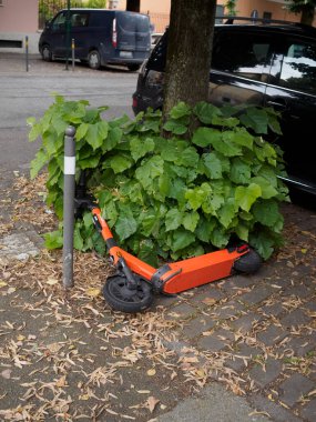 Cremona, İtalya 19 Temmuz 2024 Bir turuncu elektrikli scooter Kentin kaldırımında yeşil bir çalılığın yanında yatıyor