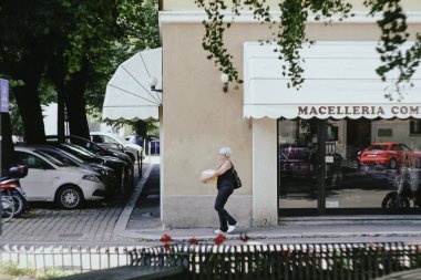 Cremona, İtalya - 15 Temmuz 2024 Kıdemli Kadın Avrupa 'daki bir kentte bir kasap dükkanının önünden geçerken bir kaldırımda yürürken erzak taşıyor. O, yiyecek için alışveriş yaptıktan sonra eve gidiyor.