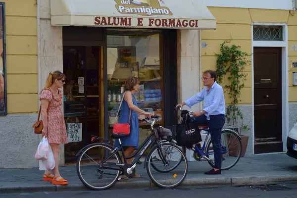 stock image Parma, Italy July 24th 2024 Three people are chatting on the sidewalk in front of a traditional italian delicatessen, holding shopping bags and riding bicycles