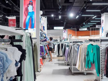 Cremona, İtalya 29 Temmuz 2024 Bir kadın moda mağazasından kıyafet alışverişi yapıyor, şık parçalar için raflara bakıyor. Modern iç mekan çeşitli yüksek kaliteli giysiler sunuyor.