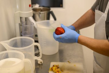 Mavi eldiven giyen biri mutfak ortamında kırmızı elma soyuyor. Arka planda çeşitli mutfak ekipmanları ve konteynırlar ve kesme tahtasındaki elma kabukları görülebilir..