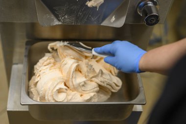 Metal bir kaptan turuncu dondurmayı kepçeleyen mavi eldiven giyen biri. Dondurma pürüzsüz bir dokuya sahiptir ve bir makineden servis edilir..