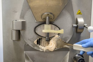 Makineden çıkarılan bir kepçeyi gösteren, çalışan bir dondurma makinesinin yakın çekimi. Makinenin, kontrol paneli ve uyarı işareti olan metalik bir kaplaması var. Kepçe kremalı ve açık renkli. Bu da yeni yapılmış dondurmayı gösteriyor..