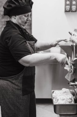 Siyah üniformalı ve şapkalı bir şef bir dondurma makinesini çalıştırıyor, mutfak ortamında taze dondurma hazırlıyor. Şef makinenin altındaki metal bir konteynırda dondurmayla göreve odaklanmış..