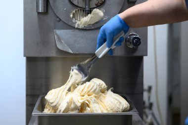 Mavi eldiven giyen biri, bir makineden kremalı dondurma almak için spatula kullanıyor. Dondurmanın pürüzsüz bir dokusu vardır ve metal bir tepside sergilenir..