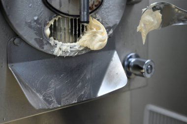 Vanilyalı dondurma dağıtan bir dondurma makinesinin yakın plan çekimi. Makinenin bir başlığı ve dondurmayı kepçeleyen bir spatulası olan metalik bir kaplaması var. Arkaplan bulanık, makinenin detaylarını vurguluyor.