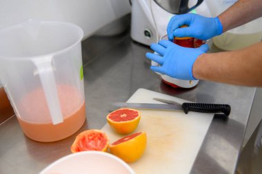 Mavi eldiven giyen biri bir kabın içine greyfurt suyu sıkıyor. Dilimlenmiş greyfurtlar bir bıçakla kesme tahtasının üzerinde. Yakında bir sürahi meyve suyu var..