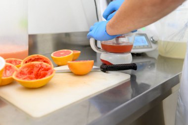 Mavi eldiven giyen bir kişi meyve karışımı hazırlamak için bir blender kullanıyor, kesme tahtasında yarı yarıya greyfurt var. Mutfak seti temiz ve düzenli. Taze malzemeler sergileniyor..