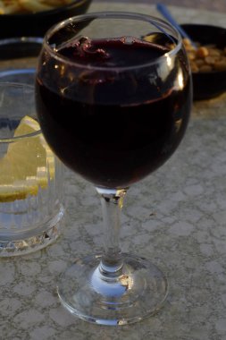 Bir bardak kırmızı şarap bir bardak buzlu ve limonlu suyla masada duruyor ve arka planda aperatifler var.