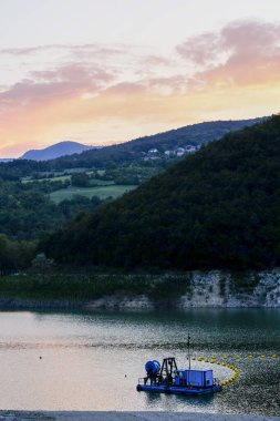 Güneş, mavi tarakla sakin gölün üzerinde batıyor dalgalar, tepeler ve ormanlar yaratıyor sıcak akşam renkleriyle yıkanmış, suya yansıyor, sakin bir manzara.