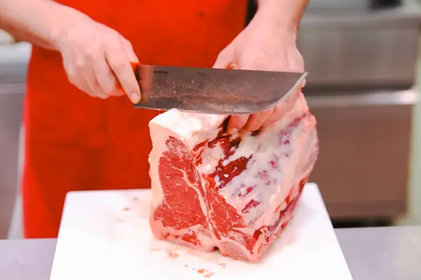 Canlı kırmızı önlüklü yetenekli bir kasap beyaz bir kesim tahtasının üzerinde taze et dilimler, hareketli bir dükkanda geleneksel zanaat sergiler.