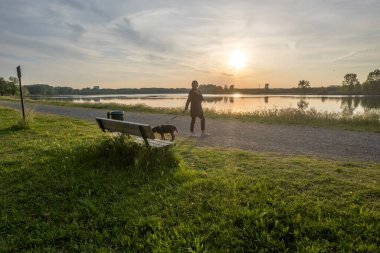 Kadın köpeğiyle huzurlu bir akşam gezintisine çıkıyor sakin bir göl kenarında, batan güneş suya sıcak bir ışık saçıyor.