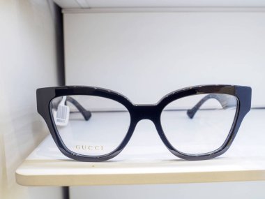 Marka isimli siyah plastik çerçeveli gözlük bir mağazanın içindeki beyaz bir rafta duruyor.