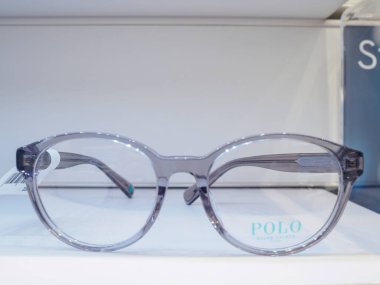 Kalın gri çerçeveli bir çift şeffaf gözlük, modern ve şık tasarımlarını sergileyen beyaz bir yüzey üzerinde sergileniyor.