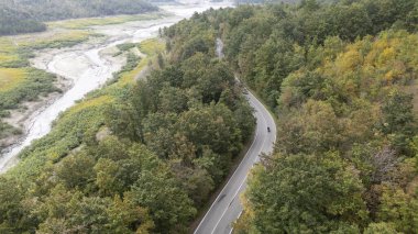 Kurumuş bir nehir yatağının yanındaki yemyeşil bir ormandan geçen dolambaçlı bir yolda giden bir motosikletlinin hava görüntüsü. Lago di Mignano, Val Tolla, PC, İtalya