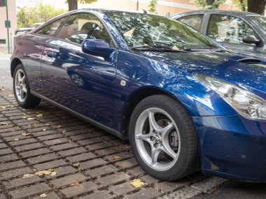 Cremona, Lombardy, İtalya 15 Ekim 2024 Tuğlalı kaldırımlı bir caddeye park etmiş parlak finalinde sonbahar ağaçlarının renklerini yansıtan parlak mavi bir Toyota Celica spor araba.