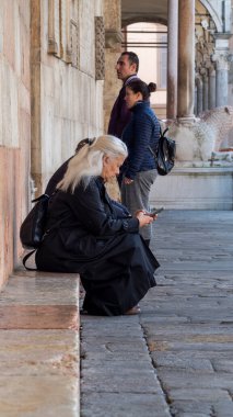 Cremona, İtalya - 3 Kasım 2024 Padua, İtalya 'da alçak bir duvarda otururken cep telefonu kullanan yaşlı kadın, yanında turistlerle birlikte