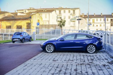 Cremona - 6 Ekim 2024 Deep Blue Metalik Tesla modeli 3 uzun menzilli, 2021, halka açık bir şarj istasyonunda pil şarj ediyor, arkasında da Renault Zoe var.