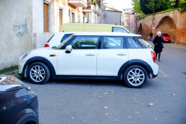 Cremona, genel olarak - 6 Ekim 2024 şık beyaz bir mini Cooper kulübü, sonbahar yapraklarıyla çevrili sakin bir yerleşim yerine zarif bir şekilde park etmiş, şehir manzarası ile kusursuz bir şekilde harmanlanmış.