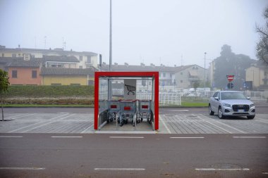 Cremona, İtalya - 7 Kasım 2024 Metal alışveriş arabaları sisli bir günde bir süpermarketin otoparkında kırmızı bir sığınağın içinde duruyor