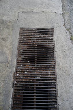 Çatlak bir kaldırıma gömülü paslı bir ızgaranın yakın görüntüsü, kuru yapraklar ve çeşitli atıklar birikmiş, kentsel çürümeyi ve bakım ihtiyacını gösteren