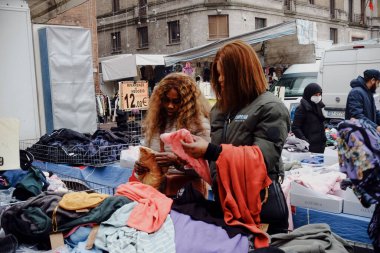 Cremona, İtalya - 24 Aralık 2024 İtalya 'da bir açık hava pazarında renkli kıyafetlere göz atan iki kadın şık kıyafetler seçiyor ve canlı atmosferin tadını çıkarıyor.