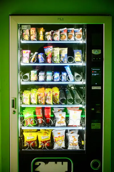 Cremona, İtalya - 30 Ocak 2025 Parlak ışıklı bir otomat çeşitli abur cubur ve içeceklerle doludur. Canlı yeşil bir duvara karşı kurulmuş. Halka açık ya da ticari bir duvar olduğunu gösteriyor.