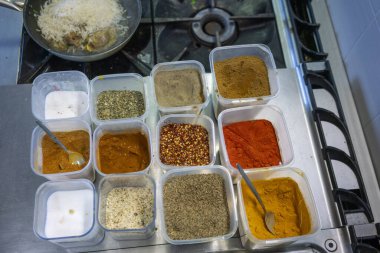 Farklı renkte baharatlar ve otlar paslanmaz çelik mutfak tezgahında küçük plastik kaplarda düzenlenirken, bir şef arka plandaki fırında peynirli bir tabak pişiriyor.