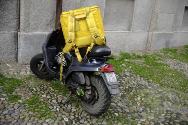 Lombardy, İtalya 1 Mart 2025 Koyu gri Yamaha Aerox motosikleti sarı izotermal yiyecek teslimatı sırt çantası ve miğferi, İtalya 'nın Cremona kentindeki kaldırım taşı caddesinde park edilmiş.