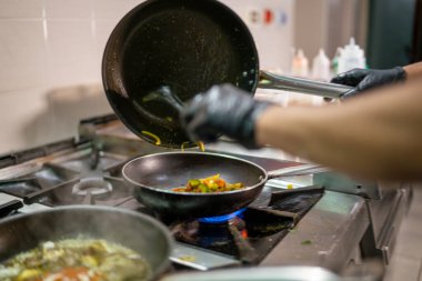 Profesyonel aşçı siyah eldiven giyer, bir tavadan diğerine Hint yemeği döker, bir restoranın mutfağında gaz sobasıyla sebze pişirir.