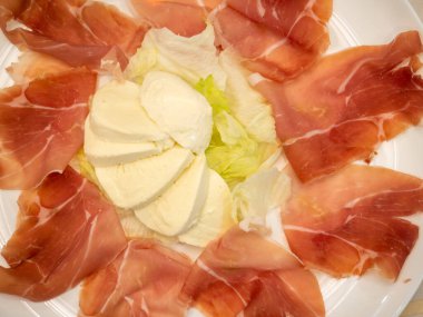 Ortasında marul yaprakları ve ince dilimlenmiş parma jambonuyla çevrili mozzarella peyniri dilimleri olan yuvarlak beyaz bir tabak.