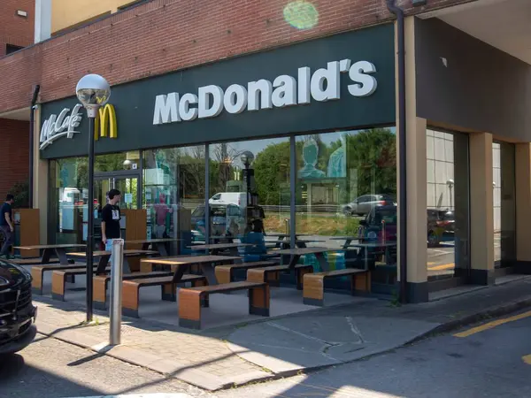 Lombardy, İtalya - 12 Mayıs 2025 İtalyan McDonald 's Sahnesi Dışarıda ahşap masa ve sandalyelerle güneşli bir gün, İnsanlar yemeğin keyfini çıkarıyor, yakınlarda arabalar park ediyor ve rahat bir atmosfer