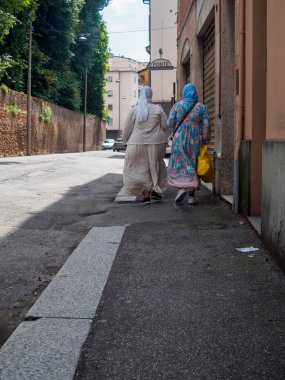 Cremona, İtalya 11 Haziran 2025. Geleneksel İslami kıyafetler giyen iki kadın, İtalyan bir kasabada dar bir sokakta ellerinde alışveriş torbalarıyla kameradan uzaklaşıyorlar.