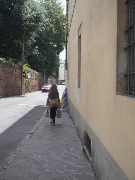 Cremona, İtalya 11 Haziran 2025 Dar Bologna caddesinde alışveriş torbaları taşıyan şık bir kadın, Müslüman kadınlar taş duvarın karşısında sohbet ediyorlar