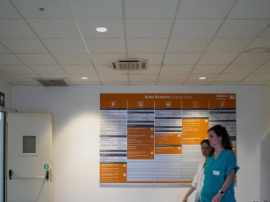 Cisanello, PI, İtalya - 13 Haziran 2025 İki hemşire farklı hastane bölümlerini gösteren turuncu bir tabelanın yanında hastane koridorunda yürüyorlar.