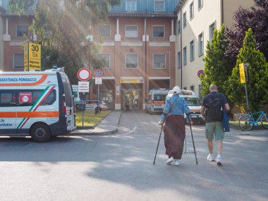 Cisanello, PI, İtalya - 13 Haziran 2025 Koltuk değneği kullanan ve bir ziyaretçinin yanında yürüyen hasta bir ambulans acil servis girişinin önünde park halindeyken, bir İtalyan hastanesine giriyor.