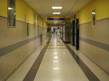 Cisanello, PI, İtalya - 13 Haziran 2025 'te uzun ve boş bir hastane koridoru, gündüz vakti bir hemşirenin acil servise girerken görüldüğü sarı ve gri renkli mimariyi gösteriyor.