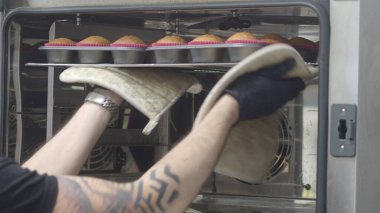 Siyah eldiven giyen profesyonel fırıncı, ticari bir fırından bir tepsi taze pişmiş keki dikkatlice kaldırıyor. Mutfak sanatında yetenek ve kendini adamışlık sergiliyor.