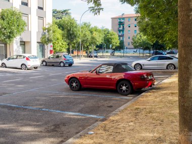 Bologna, İtalya 3 Temmuz 2025 Klasik kırmızı Mazda MX 5 Miata NA, güneşli bir yaz gününde modern arabalarla çevrili bir otoparkta park halinde, şehir planına karşı klasik tasarımın çekiciliğini sergiliyor.