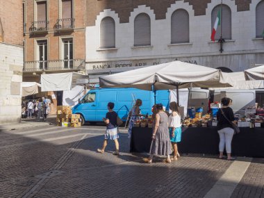 Cremona, İtalya 28 Haziran 2025 Kadınlar, güneşli bir günde Cremona, İtalya 'da bir açık hava pazarında alışveriş yapıyor, tezgahlar çeşitli ürünler satıyor ve arka planda Banca Popolare di Cremona binası bulunuyor.