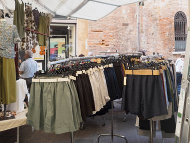 Cremona, İtalya 28 Haziran 2025 Cremona, İtalya 'da bir açık hava pazarında askıda asılı duran giysiler, arka planda bazı eşyaları düzenleyen bir satıcı ve tuğla bina