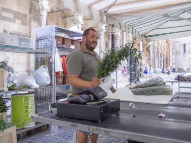 Cremona, İtalya - 15 Temmuz 2025 Gülümseyen Satıcı İtalya 'daki Bustling Outdoor Market Stall' da Taze Yeşillik ve Bitkileri Gösteriyor