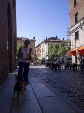Cremona, İtalya - 27 Temmuz 2024 Güneşli bir kaldırım taşı caddesinde küçük bir köpeği gezdiren bir kadın kuzey İtalya 'da tarihi binalar ve açık hava kafeleriyle dolu büyüleyici bir Avrupa seyahat sahnesi