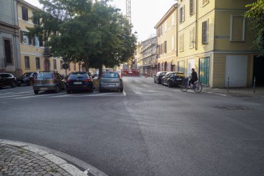Cremona, İtalya. 6 Ağustos 2025. Bisikletçi, İtalya 'nın Alessandria şehrinde güneşli bir yerleşim bölgesinde, park halindeki arabaları ve yakındaki bir inşaat sahasında asfalt bir sokakta sürüyor.