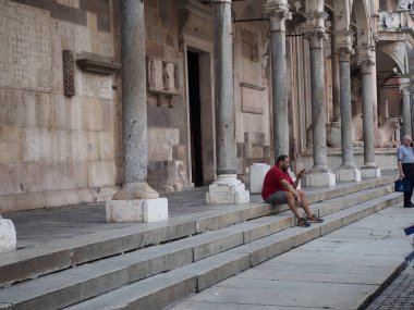 Cremona, İtalya - 6 Ağustos 2025 Adam İtalya 'da Uzun Sütunları ve Açık Kapısı olan Tarihi Taş Binanın Dışında Oturuyor