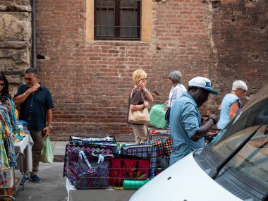 Cremona, Italy- 23 Ağustos 2025 İnsanlar Avrupa 'nın tarihi bir caddesinde sıcak İtalyan güneşinin altında el yapımı el işleri ve hediyelik eşyalar sergileyerek renkli pazar yerlerine bakıyorlar.
