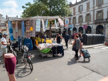 Cremona, Italy- 23 Ağustos 2025 tarihi bir İtalyan şehir meydanında güneşli bir gökyüzünün altında renkli tekstil ve giysiler sergilenen açık hava manzarası.