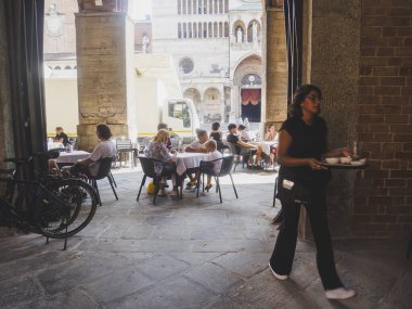 Cremona, İtalya - 3 Temmuz 2025 Kemerli porticonun altındaki açık kafe masalarında insanlar yemek yiyor ve bir garson güneşli bir günde İtalya 'nın Cremona kentindeki tarihi Duomo kompleksini net bir şekilde görüyor.