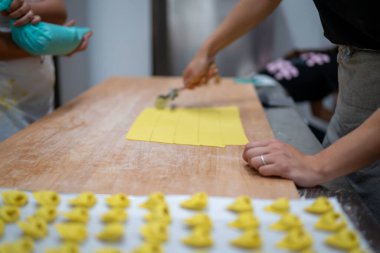 Two people are carefully making fresh homemade pasta on a large wooden table with a rolling pin creating perfect squares